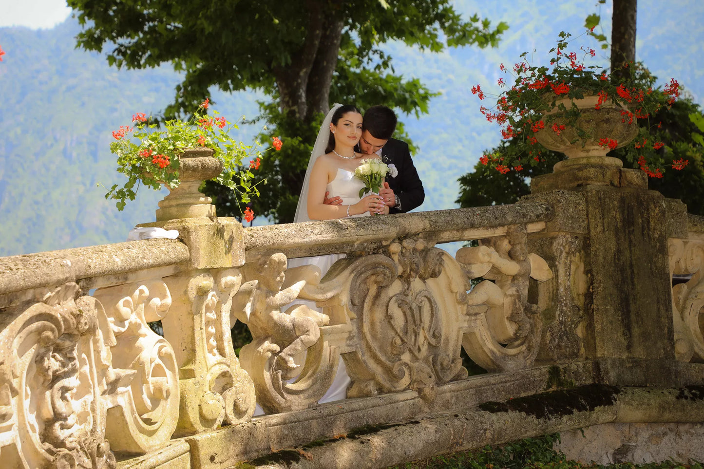 Bride and groom embracing behind the ornate stone balustrade of Villa del Balbianello with floral urns and mountain views