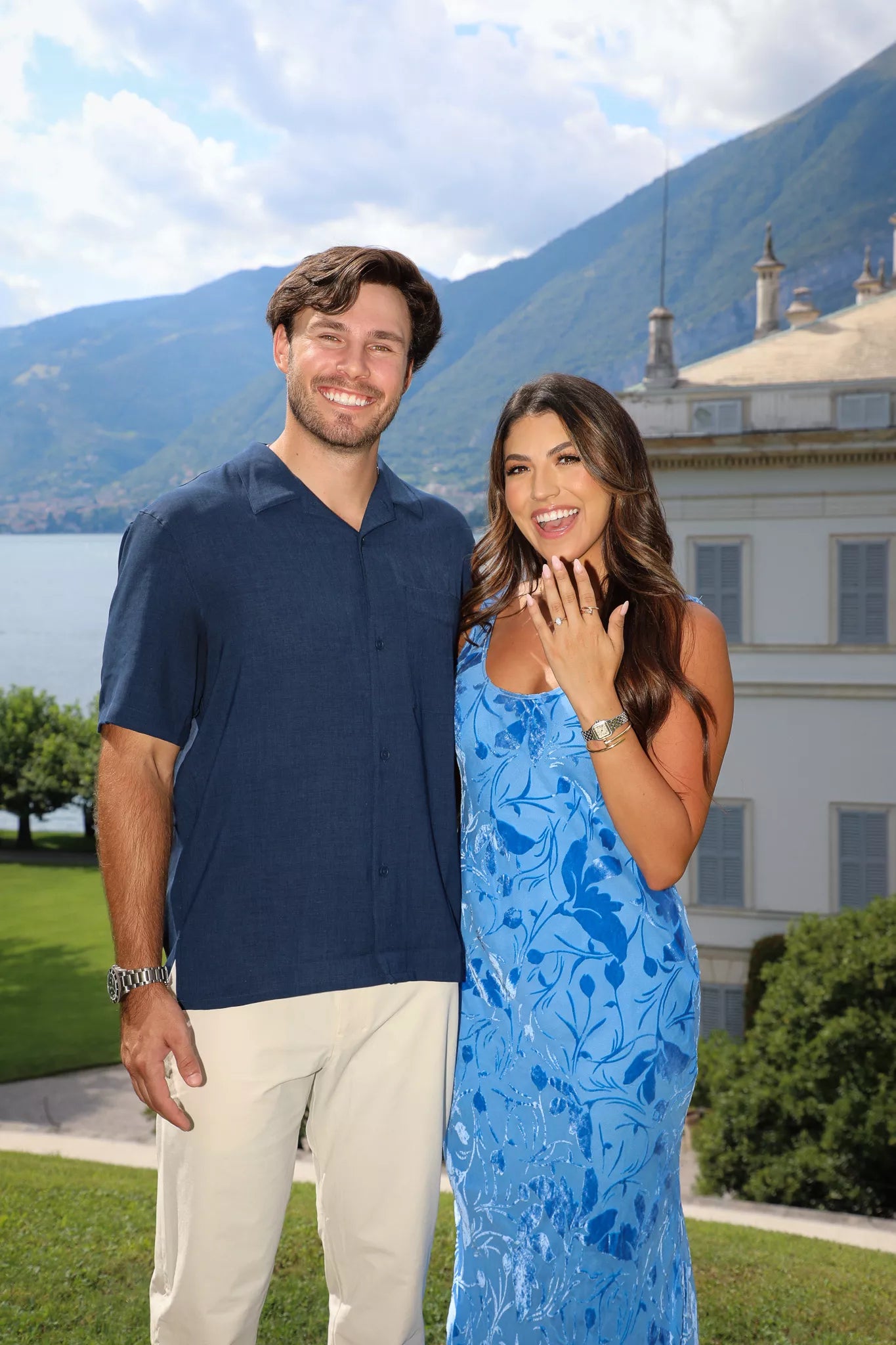Close engagement portrait of a smiling couple showing the ring with Lake Como and villa behind them