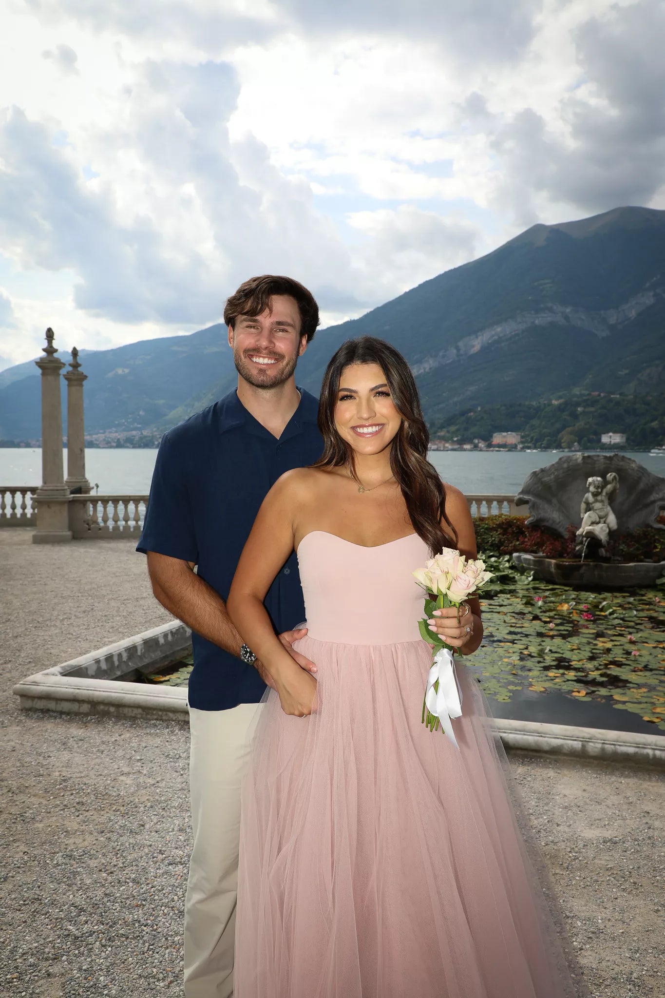 Engaged couple posing with a pink bouquet beside the lily pond and mountains at Villa Melzi in Bellagio, Lake Como