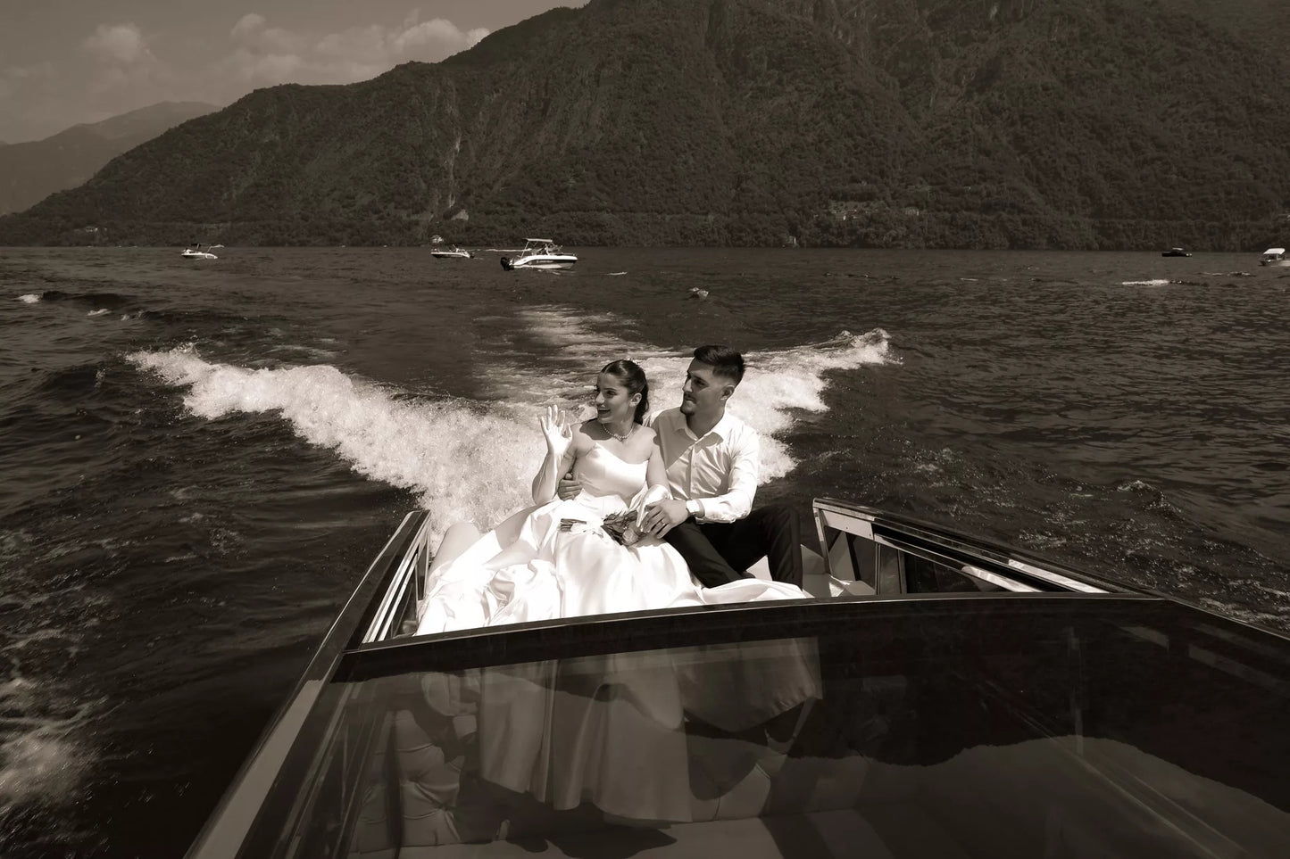 Sepia wide photo of a bride and groom cruising across Lake Como on a private boat with dramatic mountain scenery
