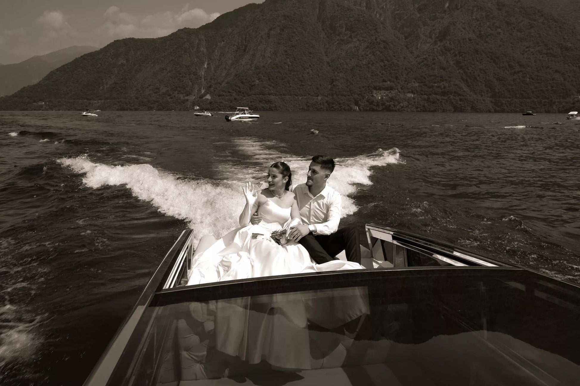 Sepia wide photo of a bride and groom cruising across Lake Como on a private boat with dramatic mountain scenery