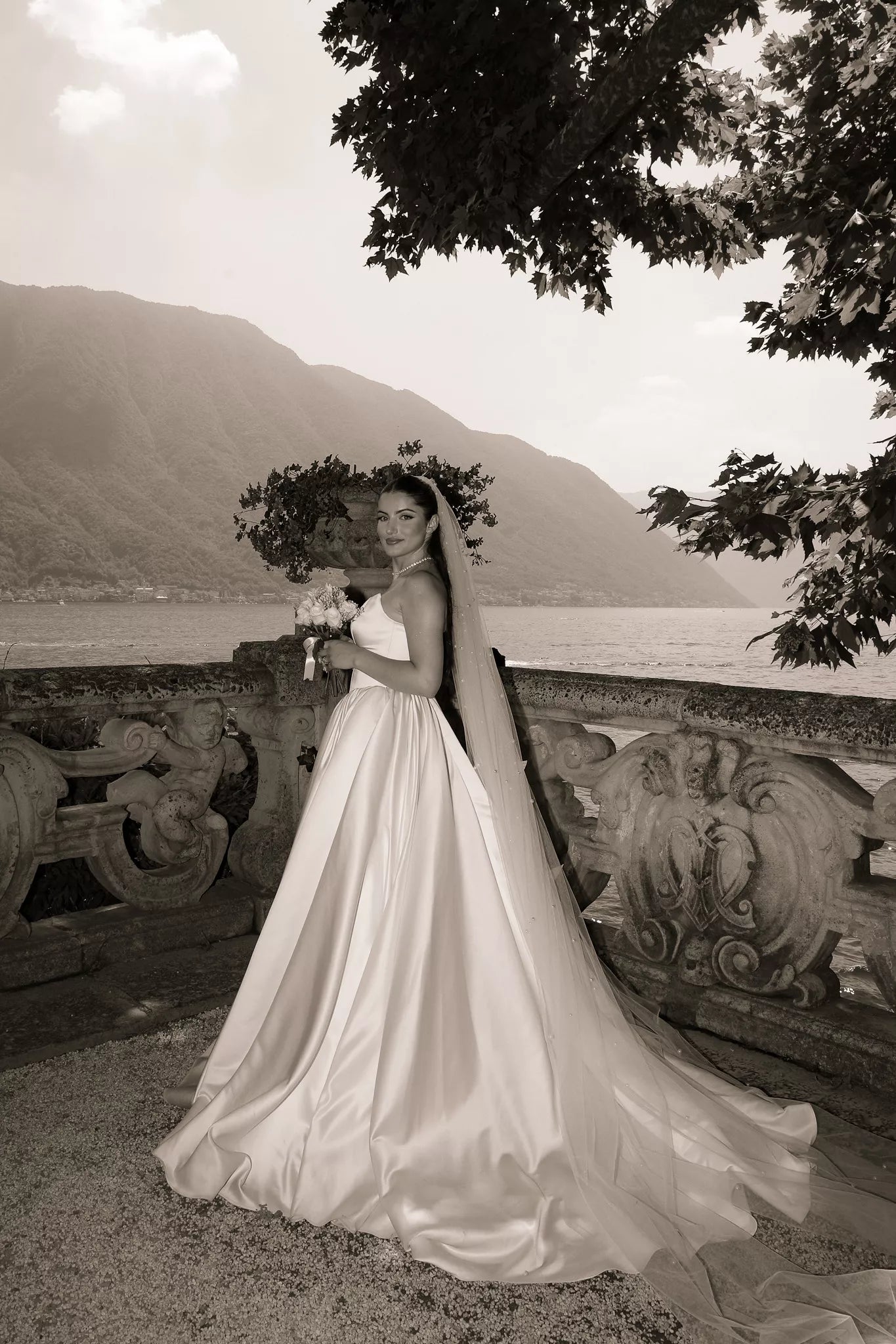 Sepia bridal portrait on a Lake Como stone terrace featuring a long veil, bouquet and mountain backdrop