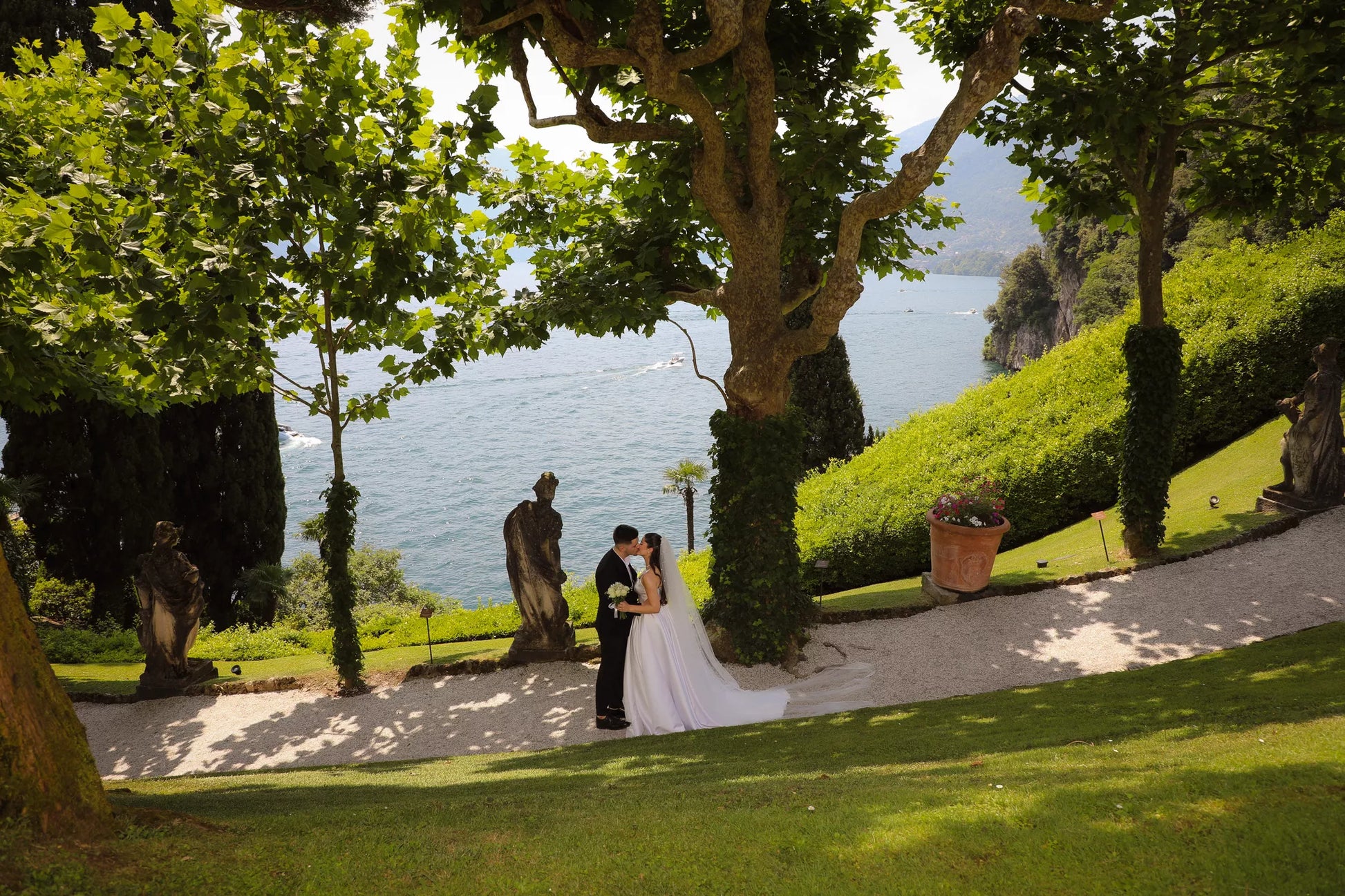 Wide garden scene of a wedding couple embracing on the hillside paths of Villa del Balbianello above Lake Como