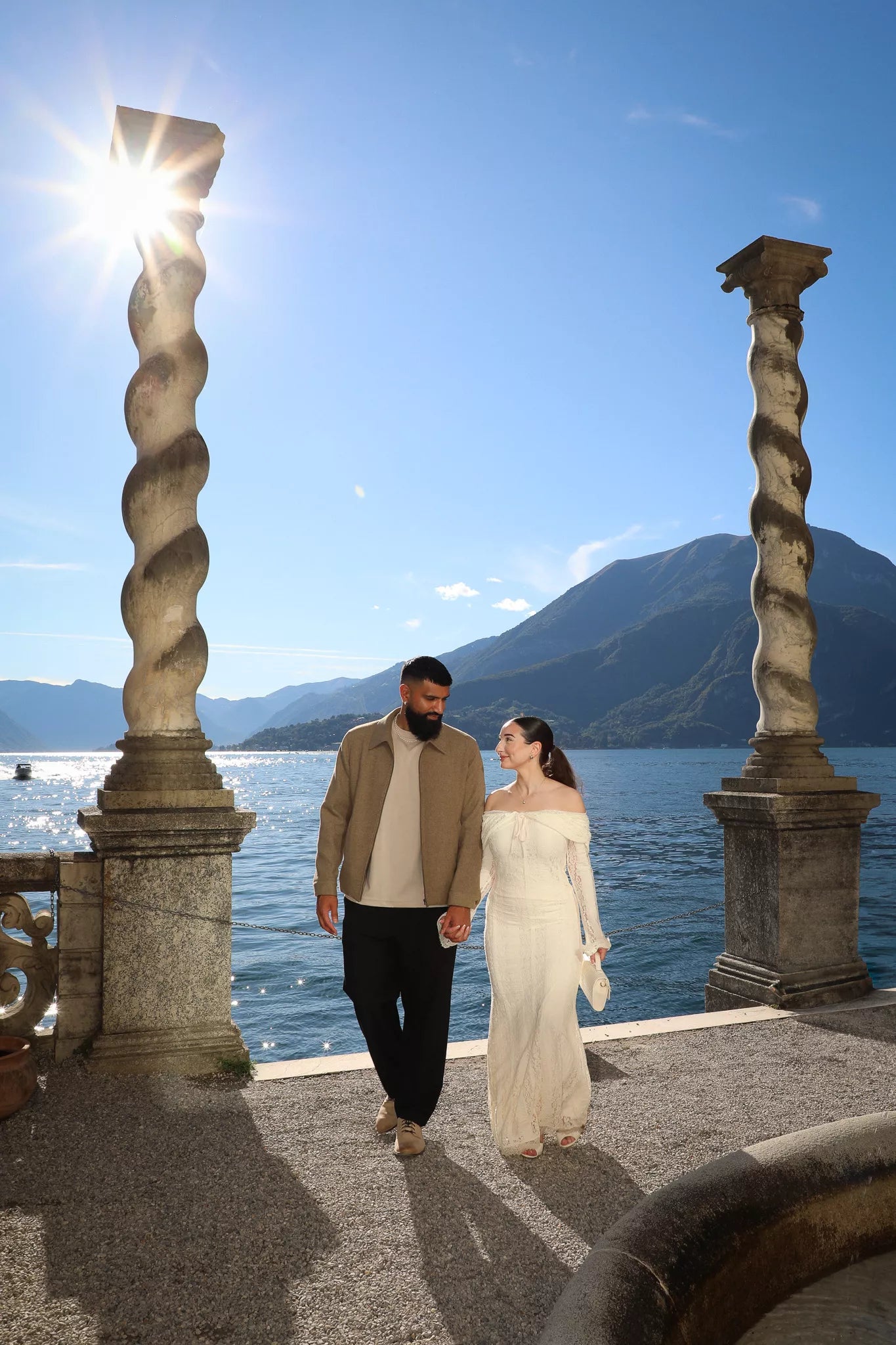 Bride in lace dress walking hand in hand with groom between stone columns overlooking Lake Como at Villa Monastero in Varenna